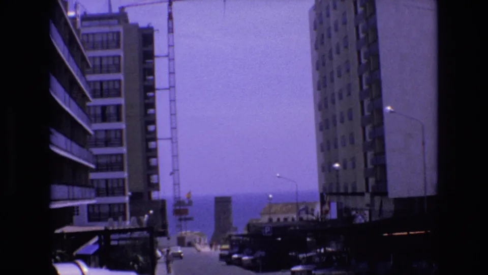 1972 Spain 8mm frame showing a purple-tinted coastal urban street between tall apartment buildings, with parked cars, small distant figures, and the sea at the end of the view.