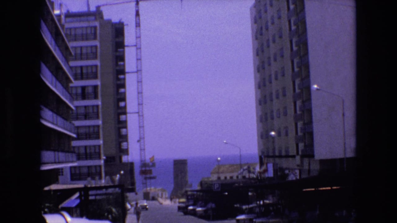 1972 Spain 8mm frame showing a coastal urban street between tall buildings, with a construction crane and distant ocean horizon.