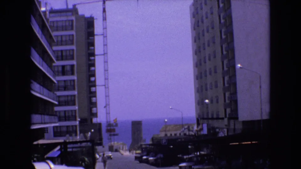1972 Spain 8mm frame showing a coastal urban street between tall buildings, with a construction crane and distant ocean horizon.