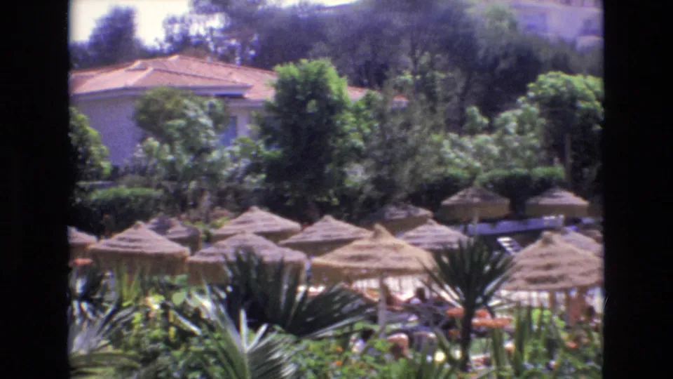 1972 Spain 8mm frame showing a resort garden with thatched huts, dense greenery, palms, and a swimming pool area.
