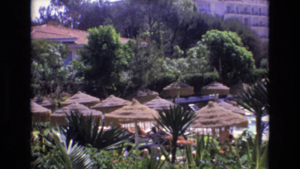 1972 Spain 8mm frame of a resort pool area with thatched shade huts, palms, dense planting, and small distant visitors.