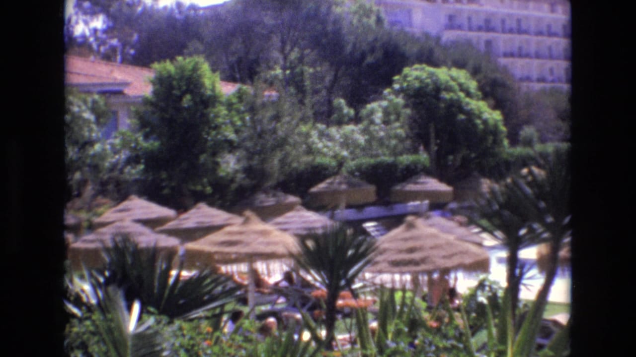 1972 Spain home-movie frame showing a resort poolside area with straw umbrellas, dense greenery, palm-like plants, lounge chairs, distant guests, and a multi-story hotel beyond the trees.