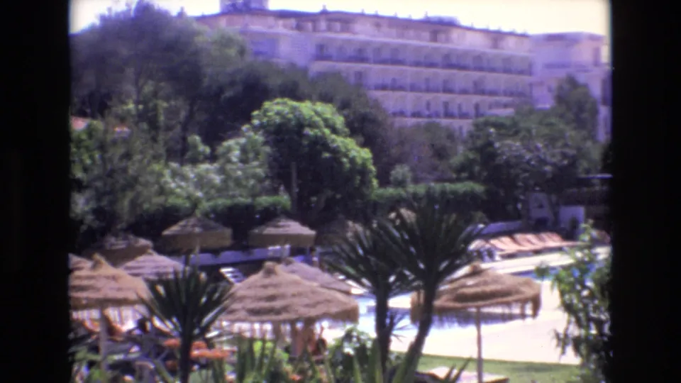 1972 Spain home-movie frame looking across straw umbrellas and palm-like plants toward lounge chairs, a bright pool area, trees, and hotel balconies in the background.