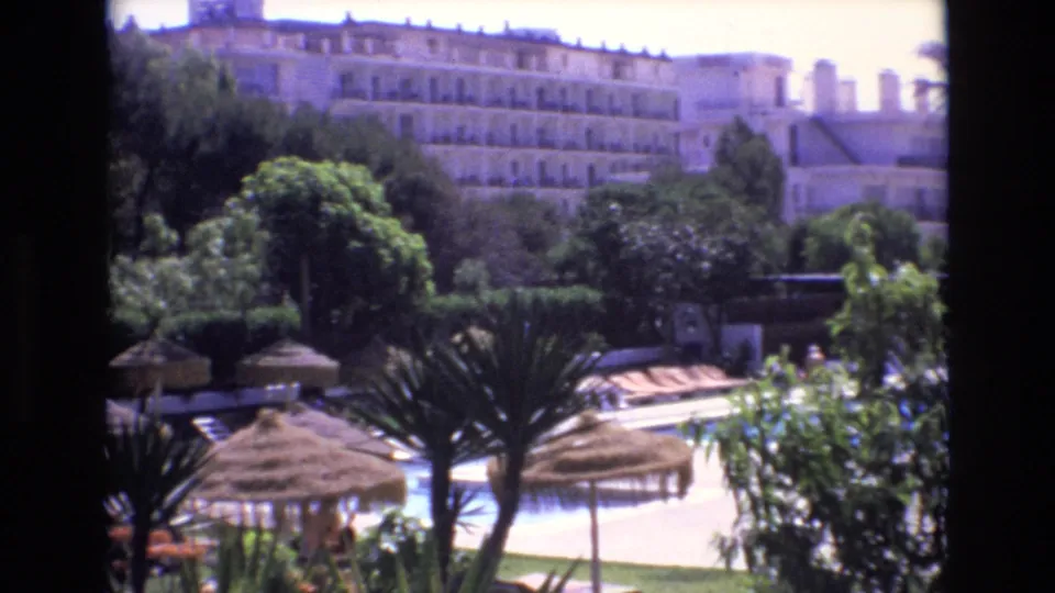 1972 Spain resort poolside scene with straw umbrellas in the foreground, dense garden planting, lounge chairs near the pool, distant guests, and a multi-story hotel behind the trees.