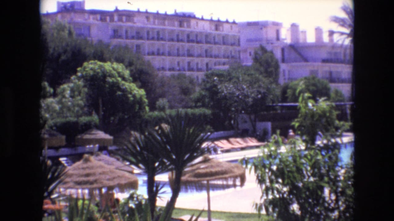 1972 Spain home-movie frame looking over a resort pool area with thatched umbrellas, palms, garden foliage, and a large multi-story hotel in the background.