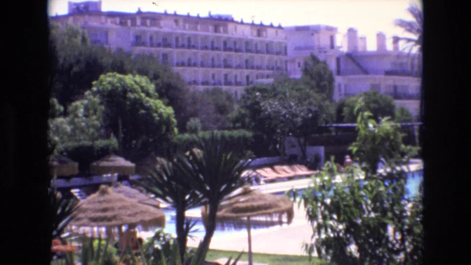 1972 Spain home-movie frame looking over a resort pool area with thatched umbrellas, palms, garden foliage, and a large multi-story hotel in the background.