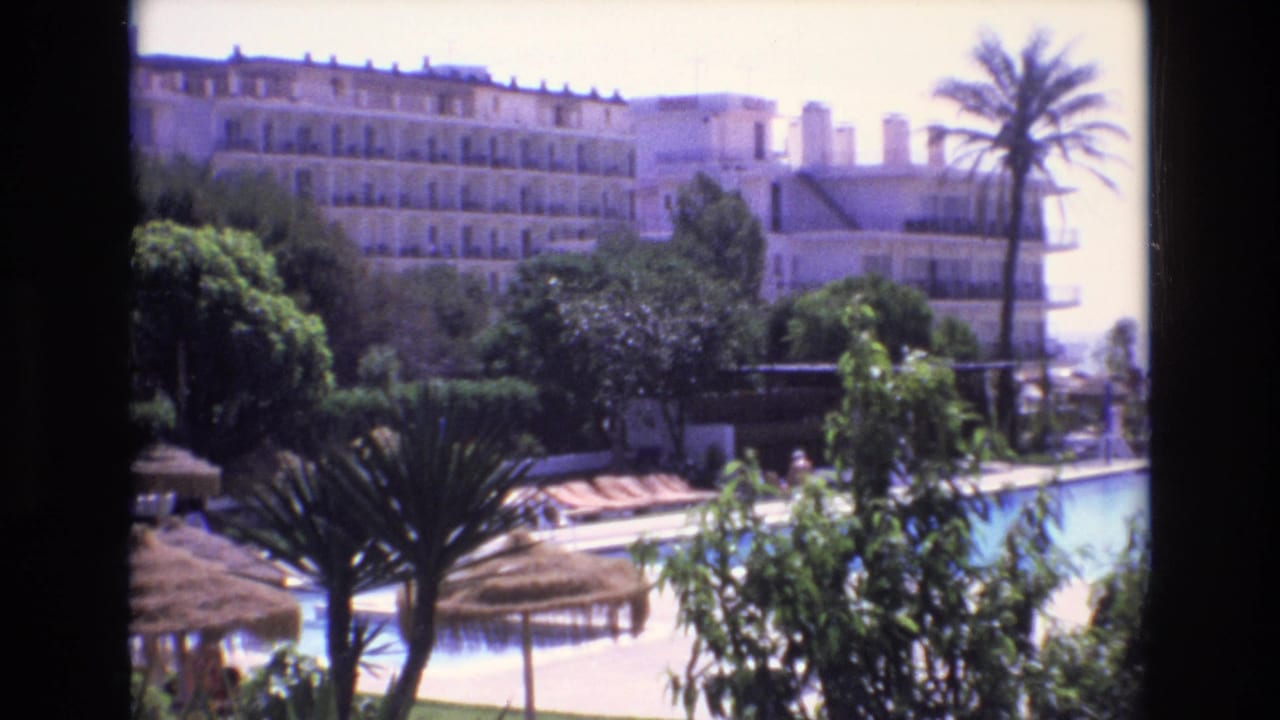 Large resort hotel behind a bright pool, palms, shrubs, and thatched umbrellas.