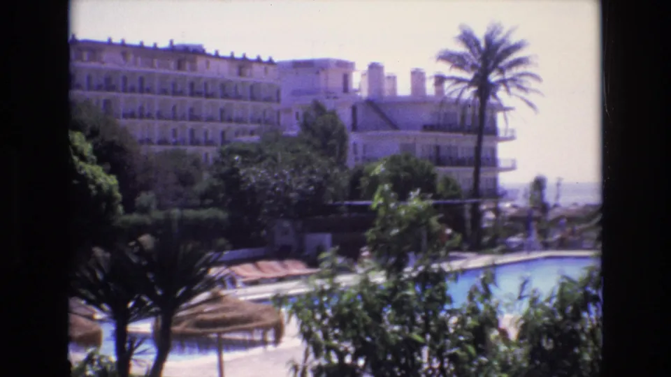 1972 Spain home-movie view across a resort pool, with palms and shrubs in front, loungers near the water, and a large hotel facing a bright coastal horizon.