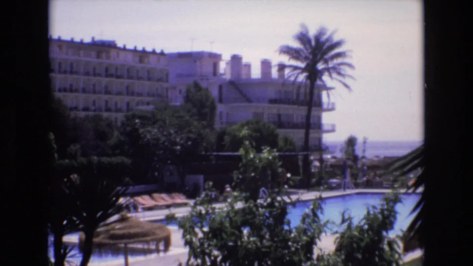 1972 Spain frame showing a resort swimming pool with a thatched umbrella, palms, foreground foliage, and a large hotel block under bright sun.