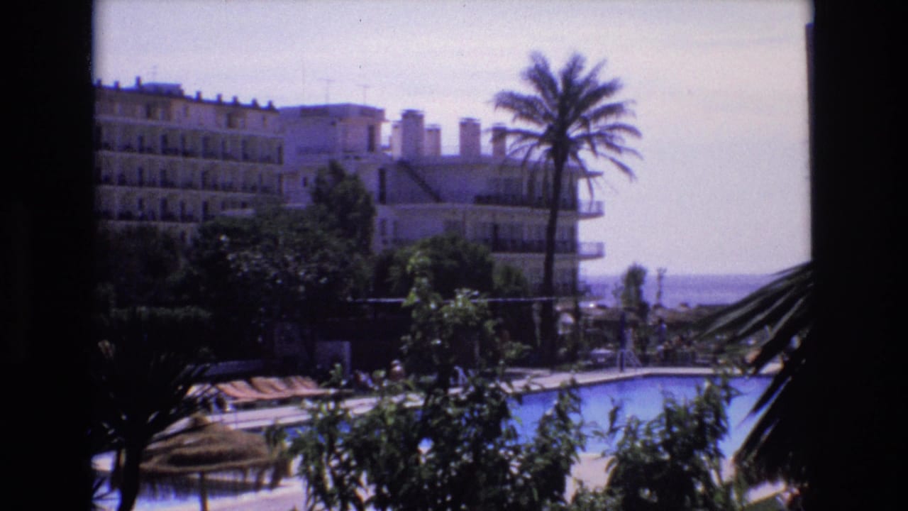 1972 Spain home-movie frame showing a swimming pool below an elevated vantage point, with lounge chairs, palm trees, white hotel buildings, and the shoreline in the distance.