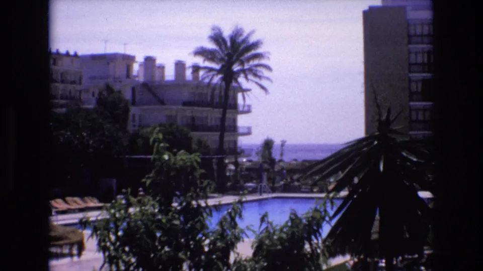 1972 Spain 8mm frame from a high vantage point, showing a resort pool terrace, rows of lounge chairs, palms, hotel structures, and the sea beyond the buildings.