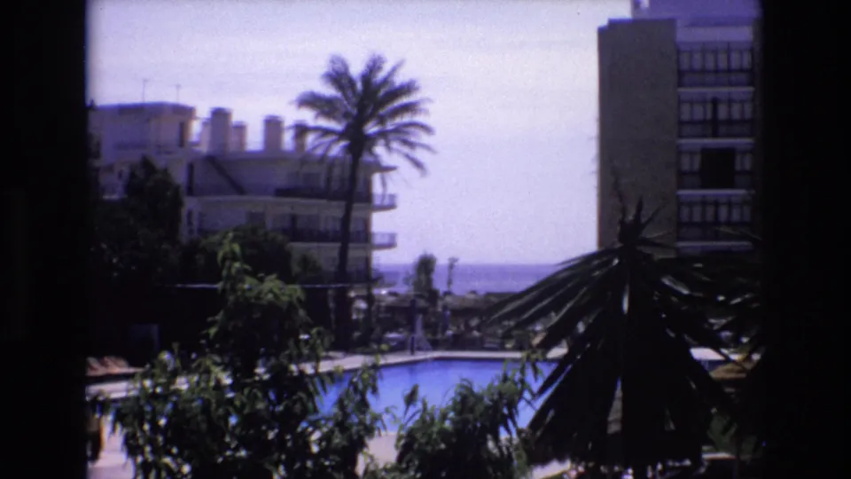 Final 1972 Spain home-movie frame showing the same elevated resort scene, with the pool partly visible below, palm silhouettes at right, hotel buildings, and the distant shoreline.