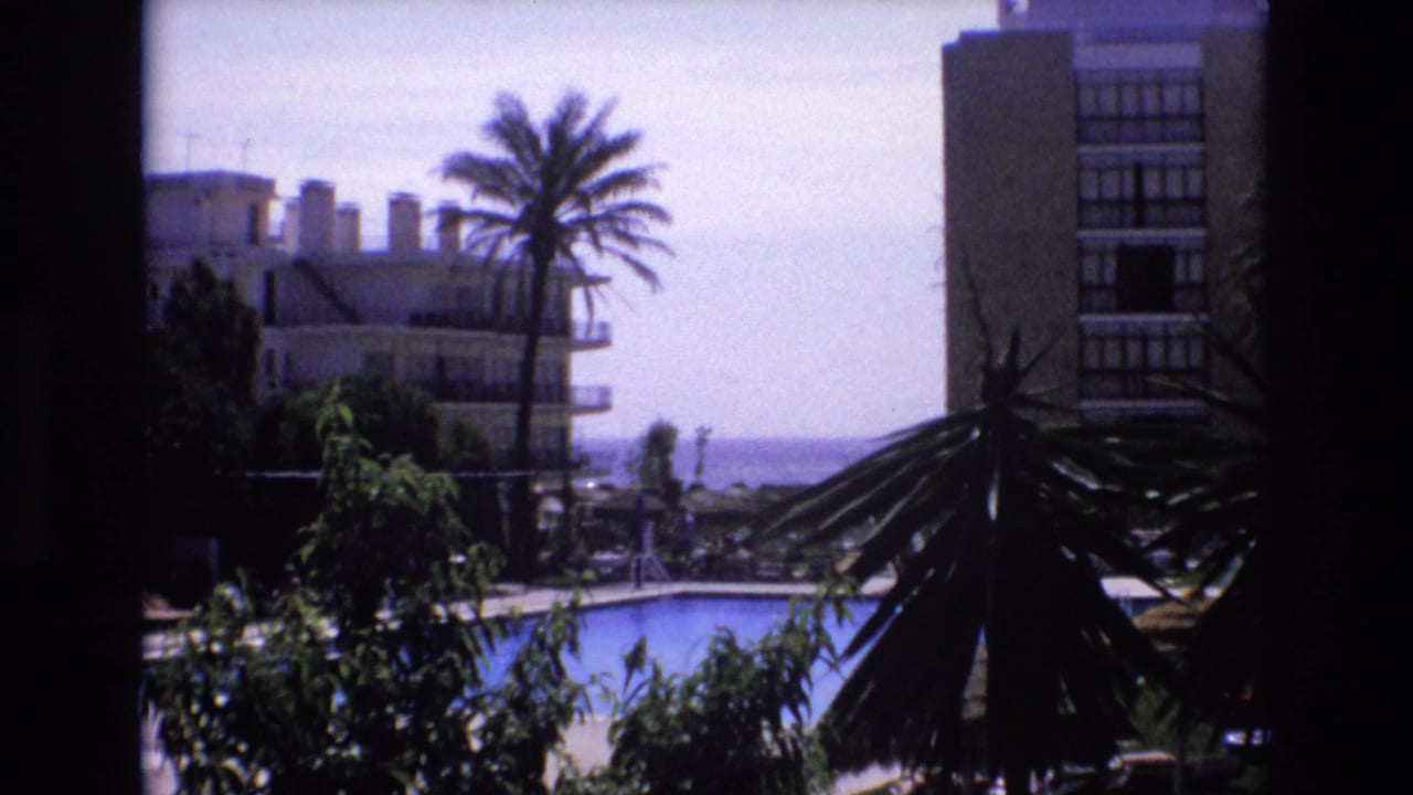 1972 Spain home-movie frame showing a resort pool area framed by foliage, with palm trees, multi-story buildings, and distant ocean visible beyond.