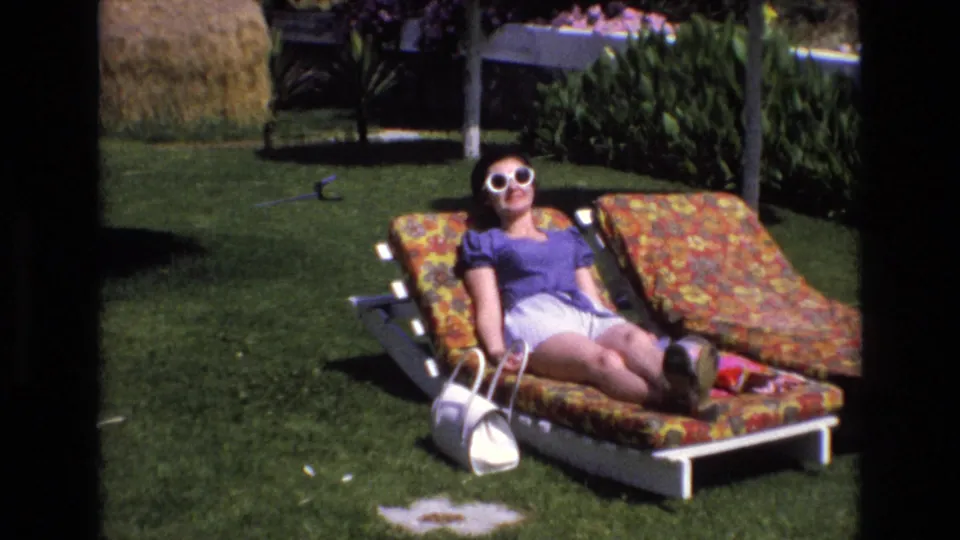 1972 Spain home-movie frame of an adult woman in white sunglasses, purple top, and white shorts sunbathing on a floral lounge chair beside a white purse.