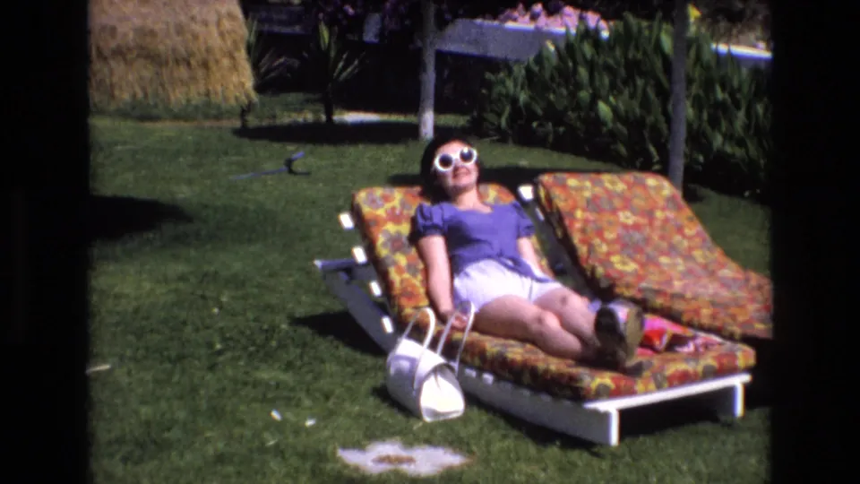 1972 Spain home-movie frame showing an adult woman relaxing on a patterned garden lounge chair, with white sunglasses, a white purse, lawn, shrubs, and a thatched shade structure visible.