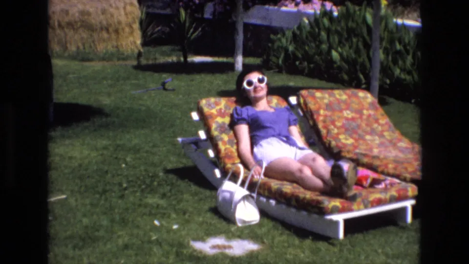 1972 Spain home-movie frame of an adult woman reclining on a floral lounge chair in a manicured garden, with a white purse, lawn, dense plantings, and bright midday light.