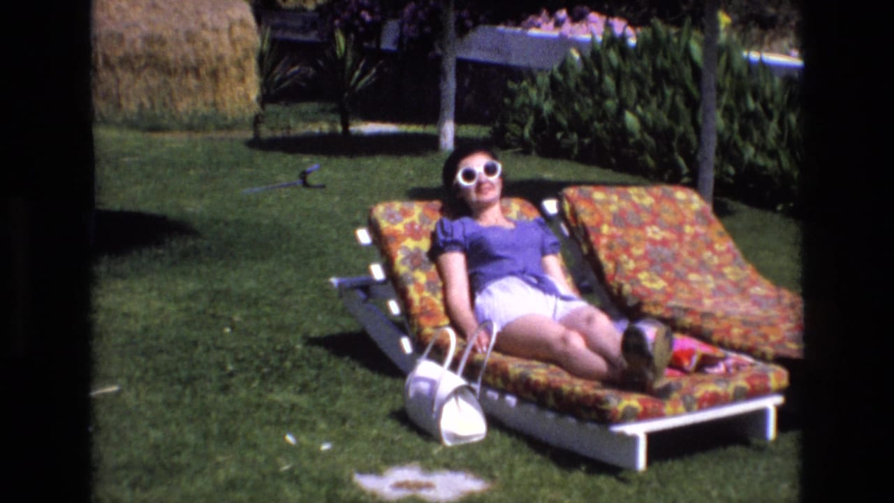 In 1972 Spain home-movie footage, a woman wearing white sunglasses, a purple top, and white shorts reclines on a floral lounge chair on a green lawn.