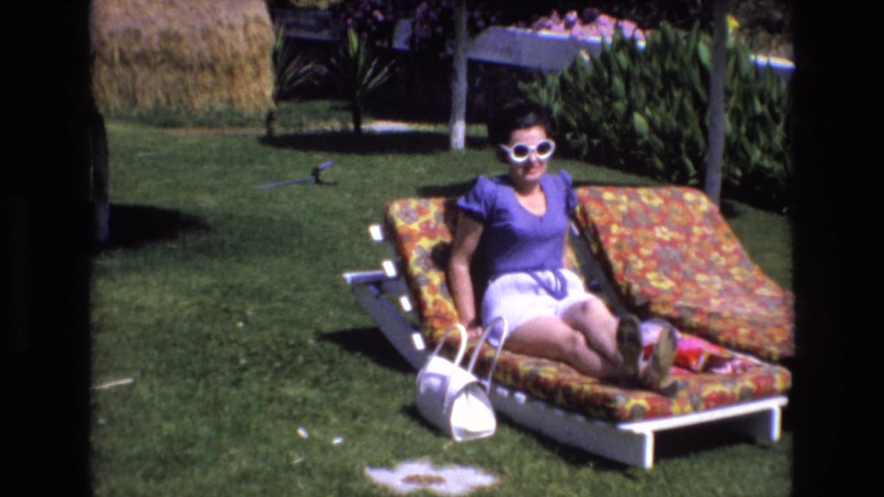 Woman in white sunglasses sits up on a floral lounge chair on a lawn with garden plants behind her.