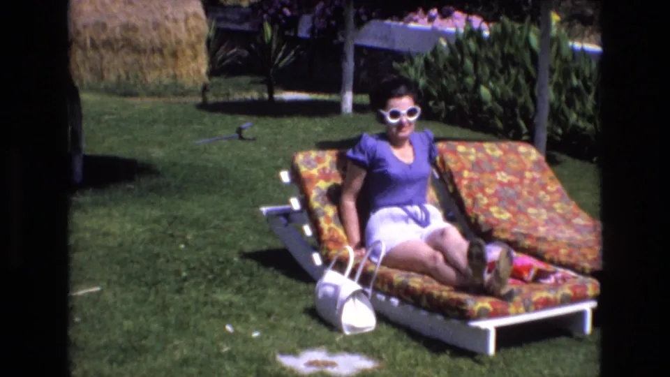 A woman in white sunglasses and summer clothes sits upright on a floral lounge chair in a sunny garden scene from 1972 Spain.