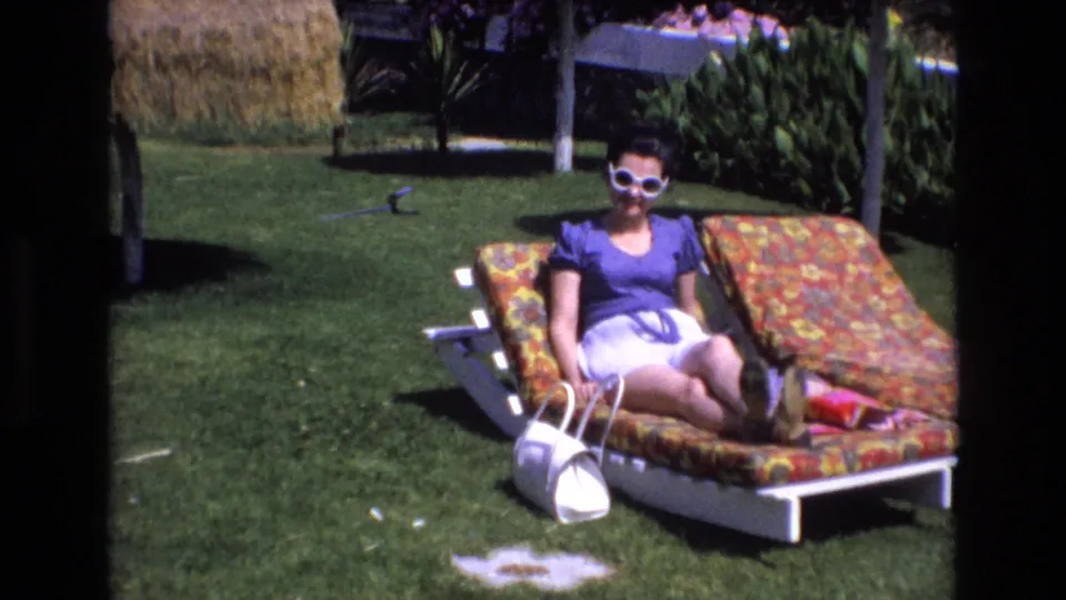 The home-movie frame shows a woman in white sunglasses seated on a floral lounge chair, with a white handbag on the grass and sunlit planting behind her.