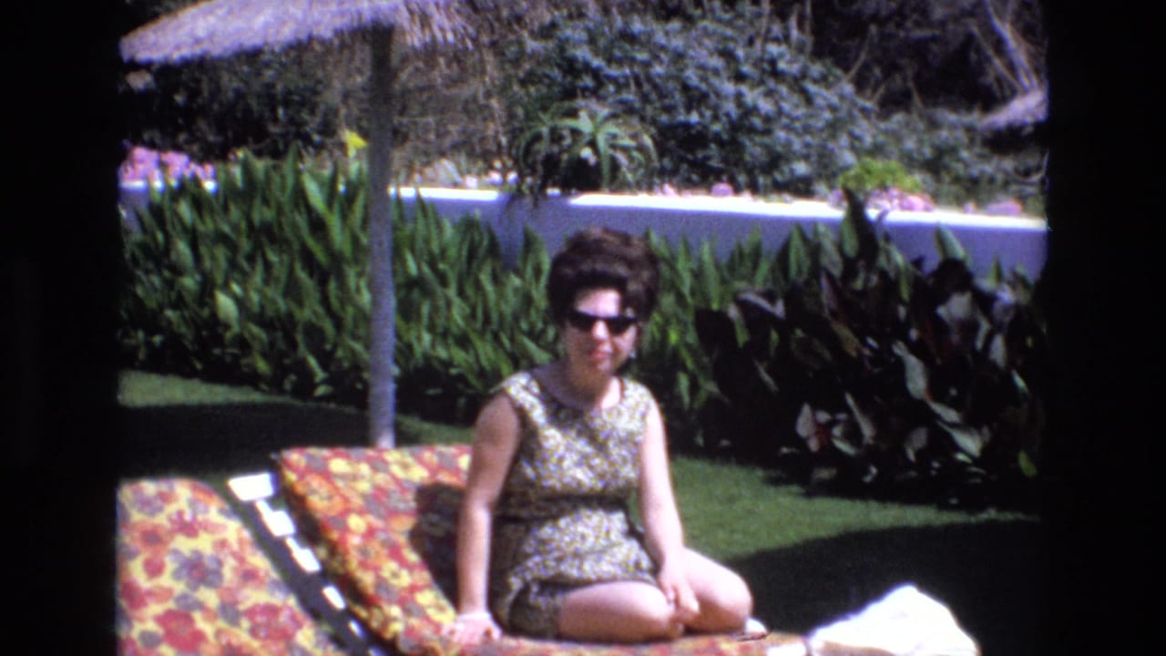 In 1972 Spain home-movie footage, a woman wearing dark sunglasses and a patterned sleeveless swimsuit sits on a floral lounge chair beside green plants and a thatched umbrella.
