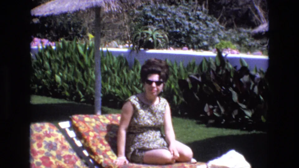 In 1972 Spain home-movie footage, a woman wearing dark sunglasses and a patterned sleeveless swimsuit sits on a floral lounge chair beside green plants and a thatched umbrella.