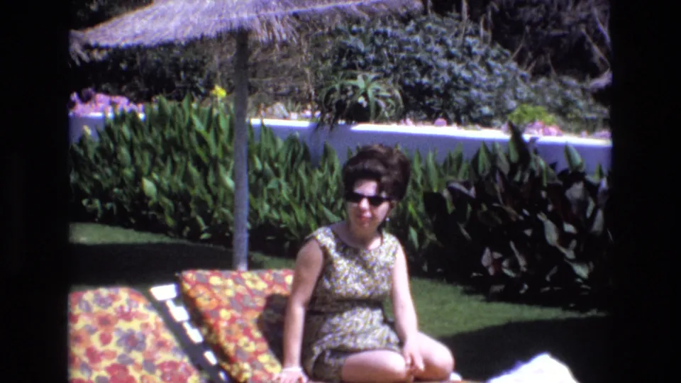 A woman in sunglasses looks toward the home-movie camera while seated on a floral sun lounger, with clipped grass, leafy plants, and a thatched umbrella visible in the 1972 Spain setting.