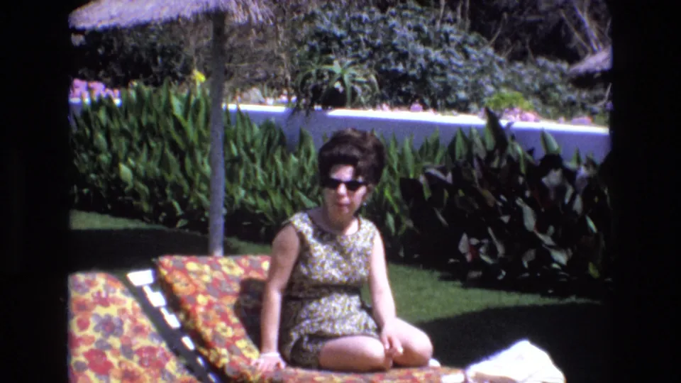Near the end of the clip, the woman sits upright on the patterned lounge chair, with her sunglasses, sleeveless swimsuit, garden foliage, lawn, and white wall still clearly visible.