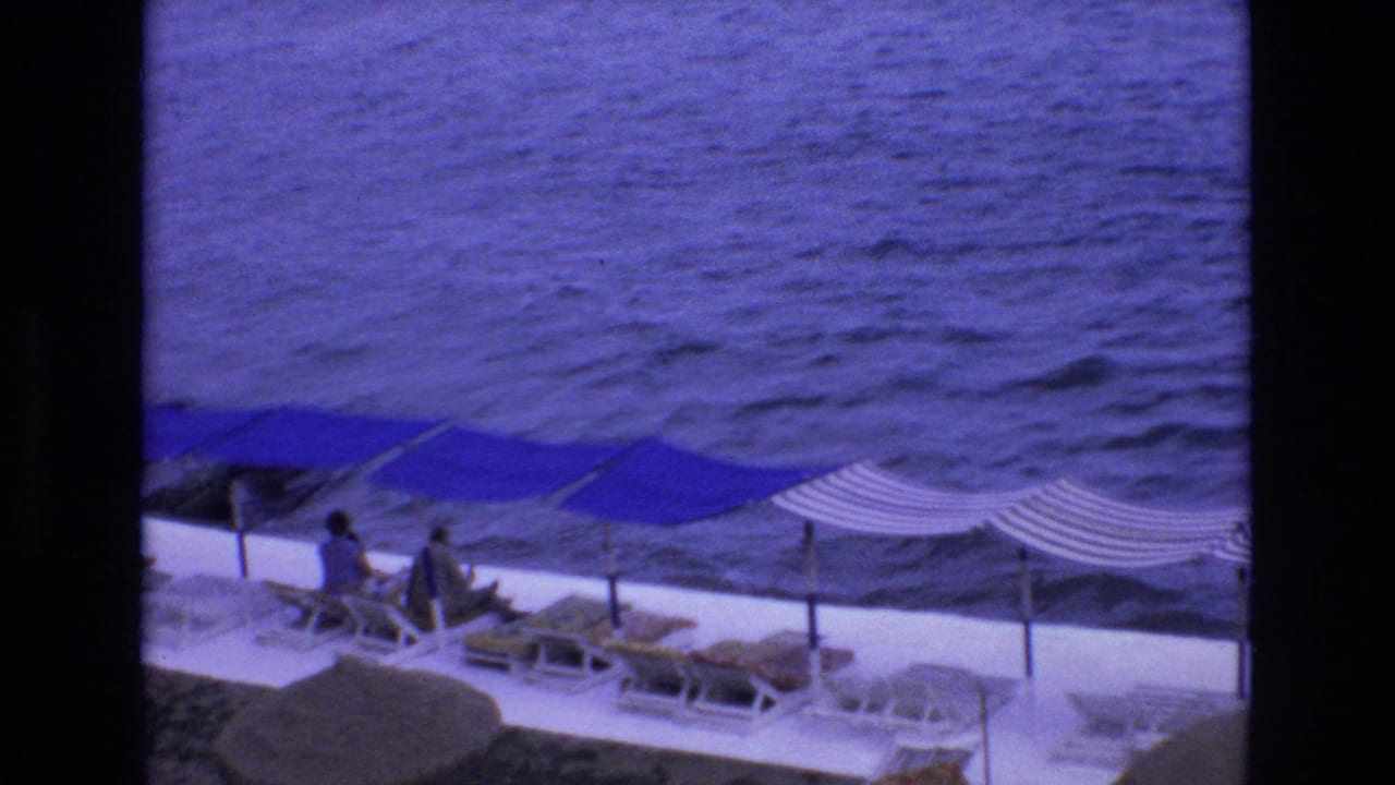 1972 Spain home-movie frame looking down at a seaside terrace with blue and striped sun canopies, lounge chairs, and a few guests beside the sea.