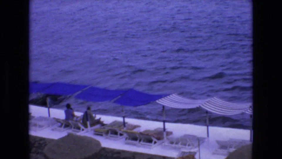 1972 Spain home-movie frame looking down at a seaside terrace with blue and striped sun canopies, lounge chairs, and a few guests beside the sea.