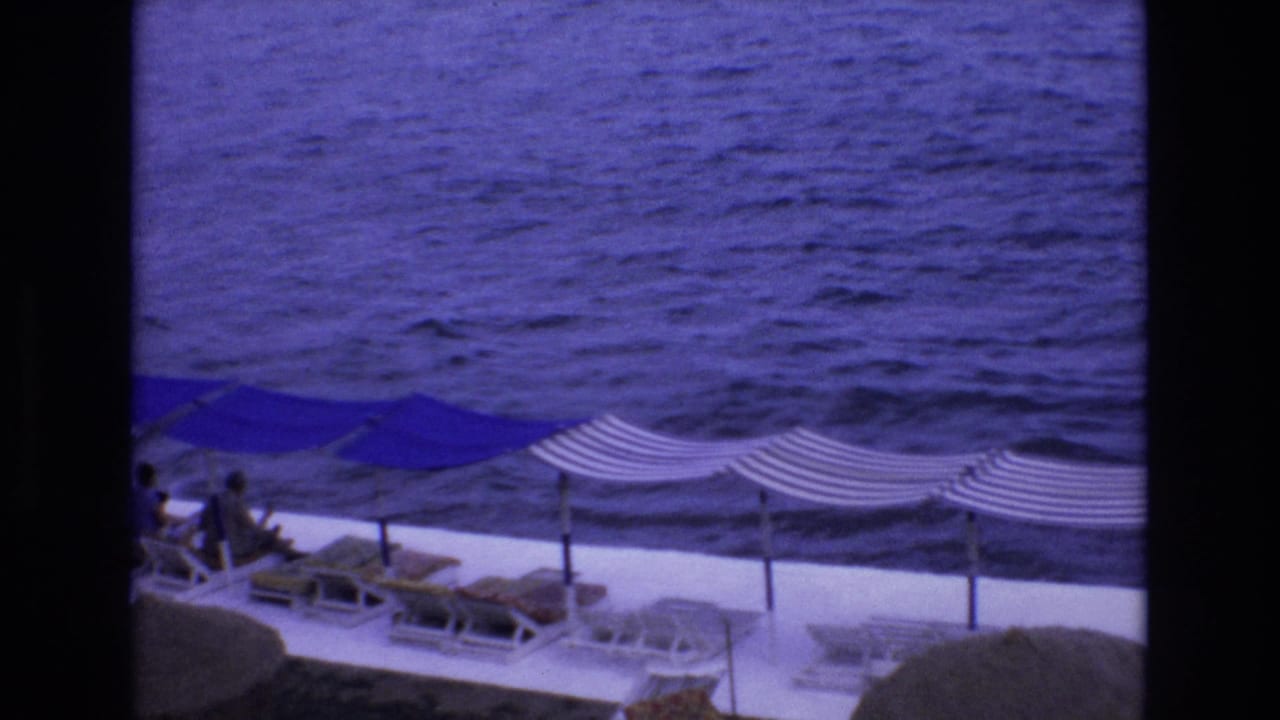 Seaside terrace with blue umbrellas, striped canopies, lounge chairs, and distant people beside choppy water.