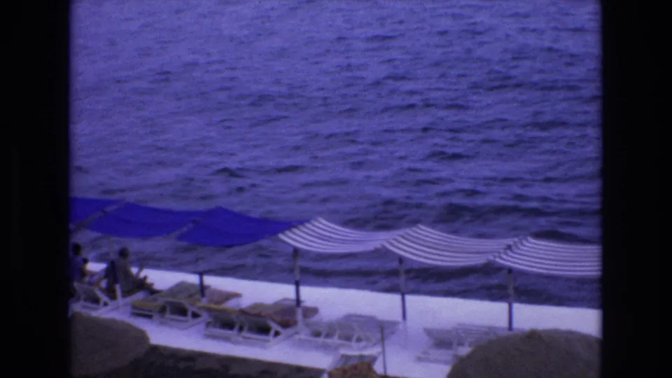 1972 Spain 8mm frame showing a high-angle resort terrace with sun canopies and lounge chairs overlooking rippled blue sea, with people visible at a distance.