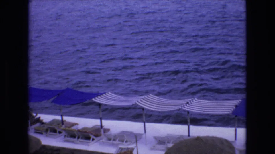 1972 Spain home-movie still with striped sun canopies and lounge chairs along the lower edge while the blue sea dominates the view.