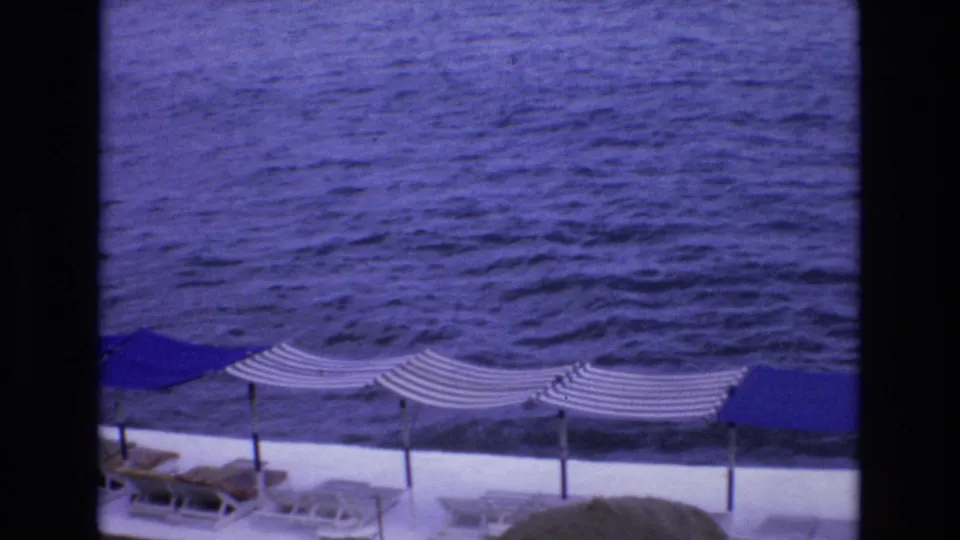 1972 Spain 8mm frame of a seaside terrace seen from above, with striped canopies, lounge chairs, a blue umbrella, and textured water beyond.