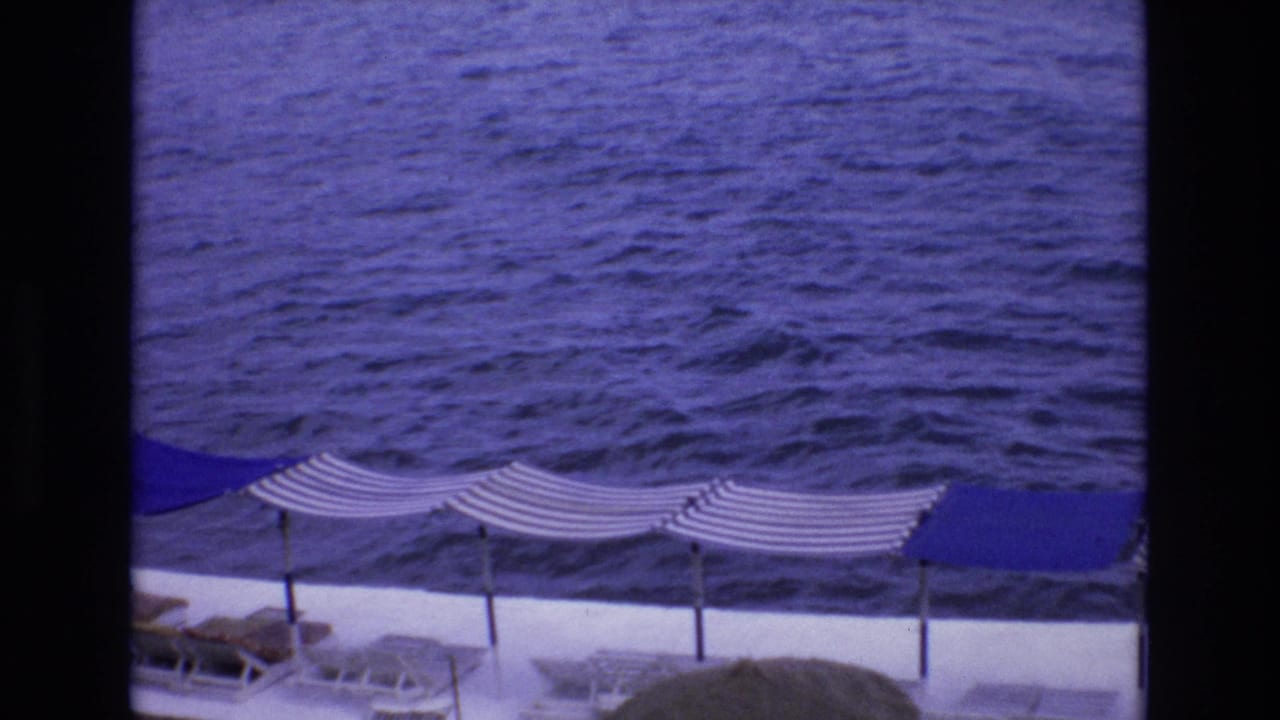 1972 Spain 8mm frame showing a blue ocean view past striped beach umbrellas, solid blue canopy sections, and lounge chairs on a white seaside deck.