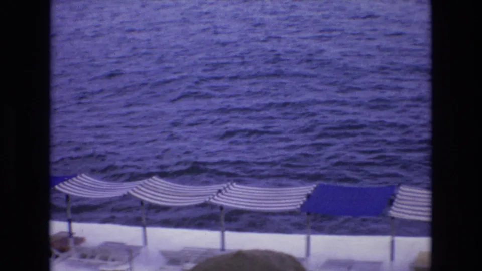 1972 Spain 8mm frame with a steady terrace view of striped beach umbrellas, a blue canopy, lounge chairs, and rippling sea water beyond.