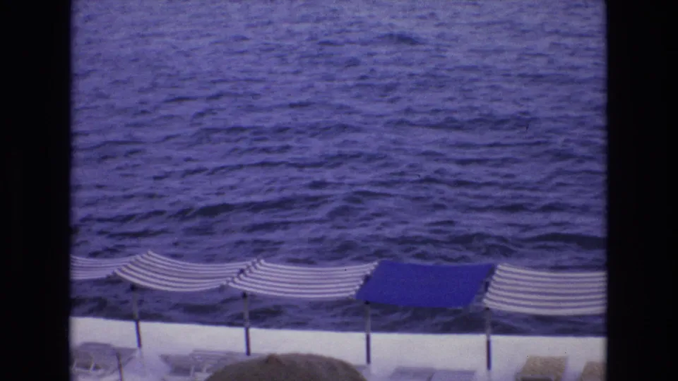 1972 Spain 8mm frame showing the ocean above a row of shaded terrace umbrellas and lounge chairs, with visible film grain and blue color shift.