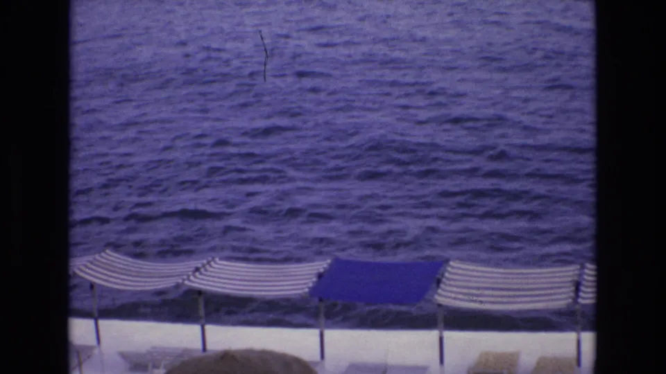 1972 Spain 8mm frame from an elevated seaside viewpoint, with rippling blue water, striped umbrellas, solid blue canopy sections, and lounge chairs on a white deck.