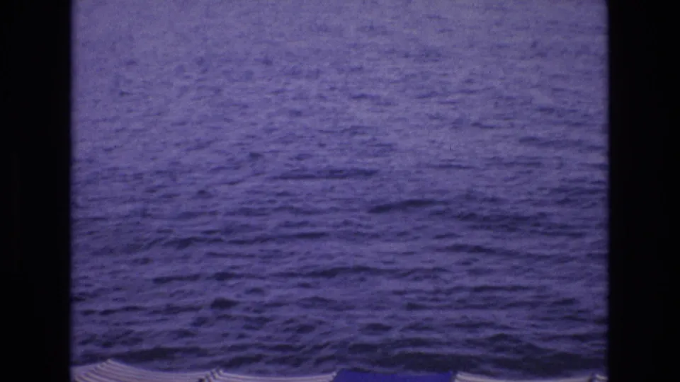 1972 Spain 8mm frame of rippling blue water, with partial striped umbrellas and a pale shoreline band at the bottom.