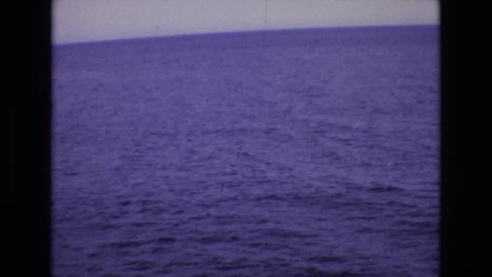1972 Spain 8mm frame showing open blue water, a pale shoreline or seawall band, and small partial umbrella shapes near the upper edge.