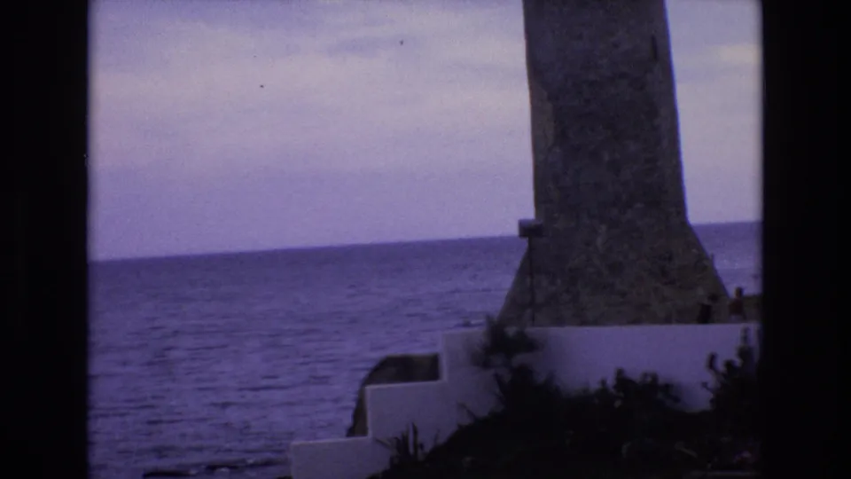 Final 1972 Spain 8mm frame showing a stone coastal tower or lighthouse-like monument near the sea, with white walls, steps, and low greenery.