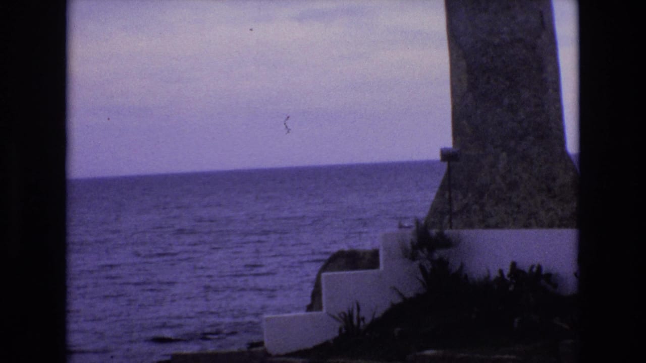 1972 Spain 8mm frame showing a purple-tinted ocean horizon, a stone lighthouse-like structure at right, and white concrete steps near dark foliage.