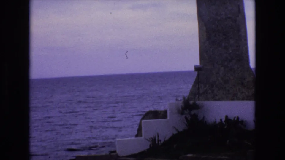 1972 Spain 8mm frame showing a purple-tinted ocean horizon, a stone lighthouse-like structure at right, and white concrete steps near dark foliage.