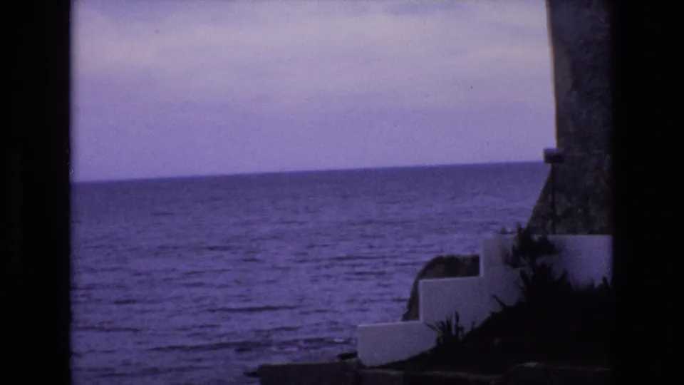 1972 Spain coastal home-movie frame with open water, a level horizon, white concrete seaside walls, dark plants, and part of a stone tower-like form at right.