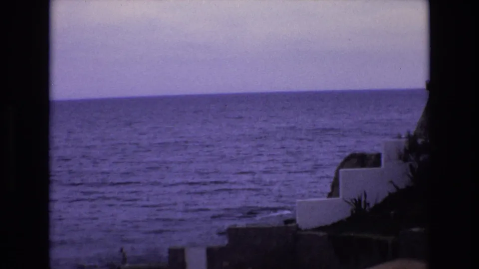 1972 Spain 8mm frame looking across the sea from a raised coastal edge, with white stepped walls, dark vegetation, and a partial stone structure at the right side.