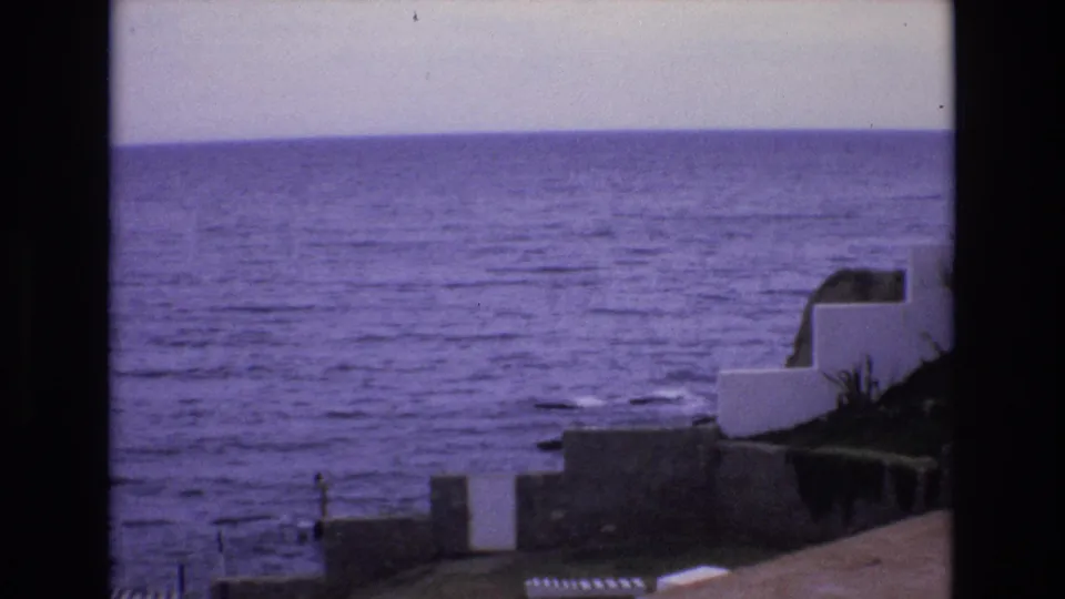 1972 Spain coastal frame showing the ocean horizon, white terrace-like walls, a sloped foreground surface, dark foliage, and low shoreline structures under a purple cast.