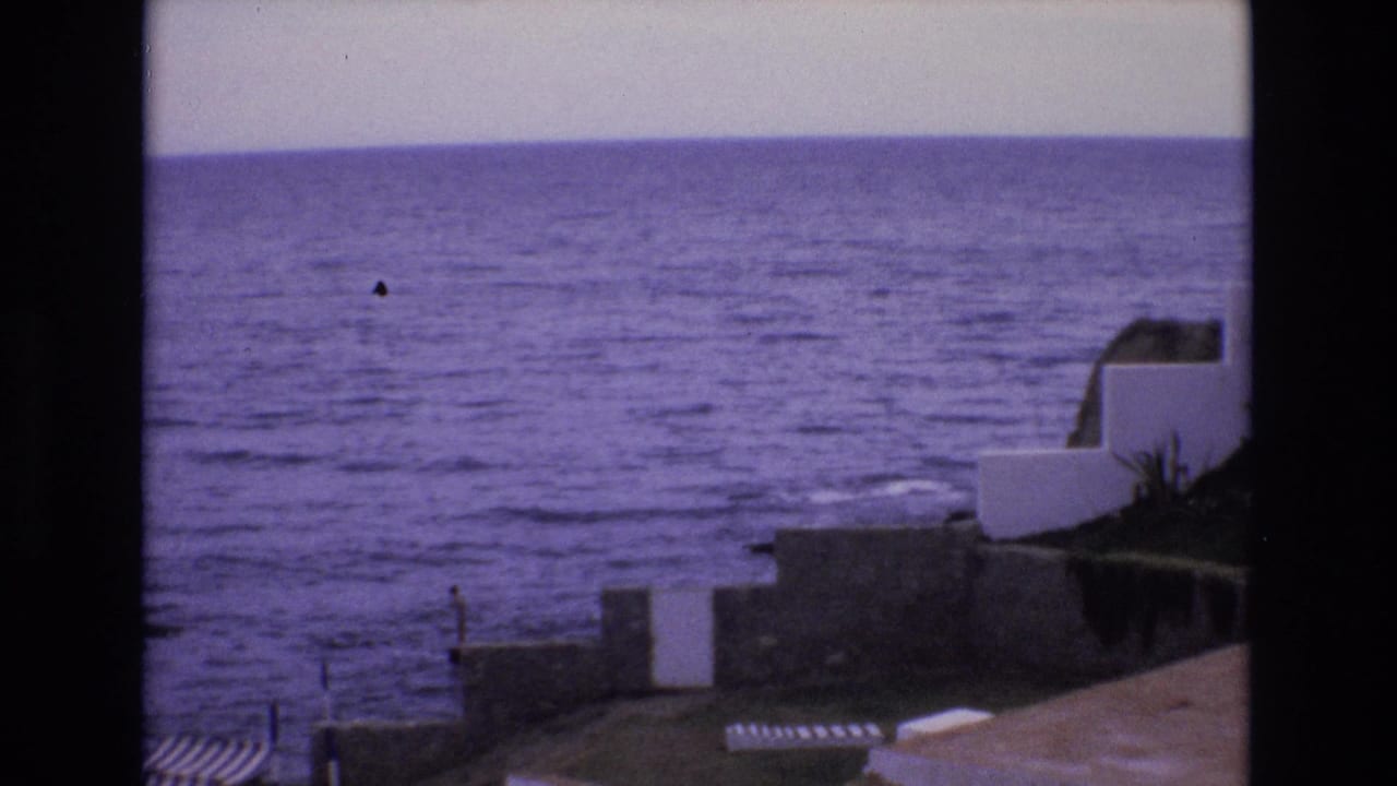 1972 Spain 8mm frame showing a purple-tinted ocean horizon, low waves, white coastal structures, stone walls, and striped beach shelters viewed from above.