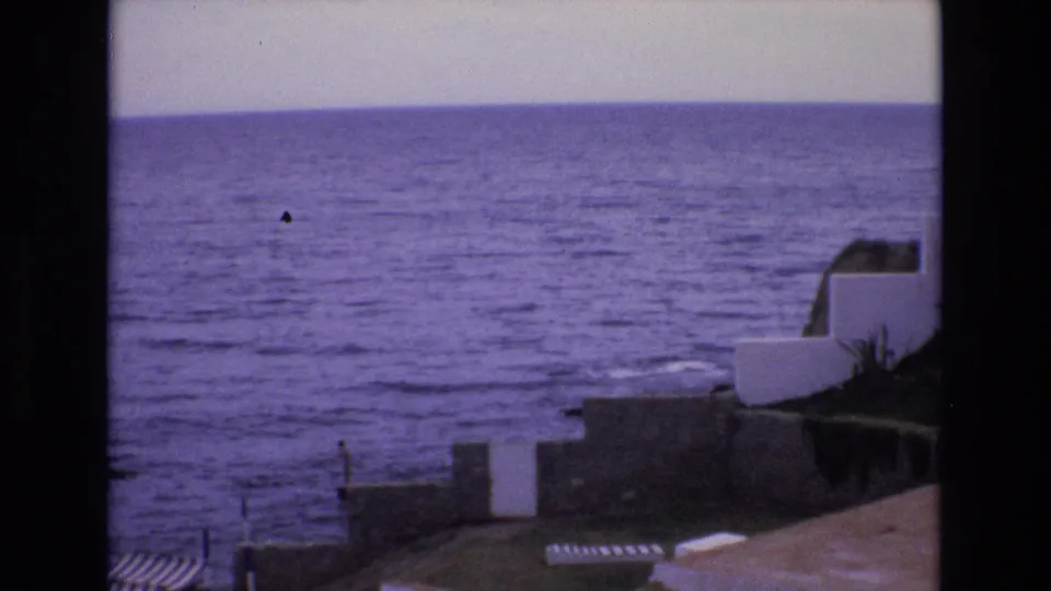 1972 Spain 8mm frame showing a purple-tinted ocean horizon, low waves, white coastal structures, stone walls, and striped beach shelters viewed from above.