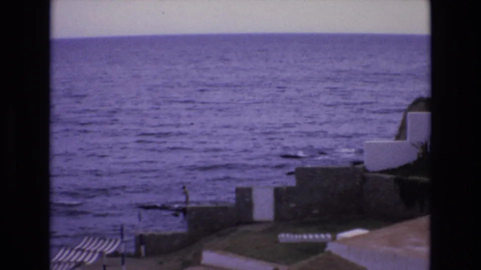 1972 Spain 8mm frame with incoming tide, stone shoreline walls, white coastal architecture, and a small person visible near the water's edge.