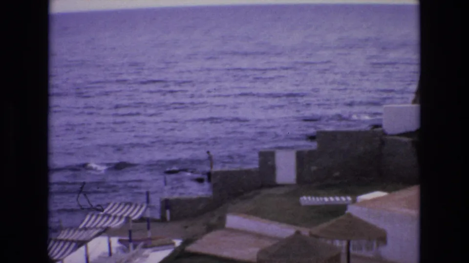 1972 Spain 8mm frame showing waves moving toward a stone waterfront below white coastal buildings and striped beach shelters.
